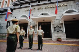 Military personnel stand outside Palacio Del Gobernador in a formal ceremony with flags displayed.