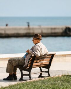 Senior man in hat sits on a bench, enjoying ocean view on a bright day.